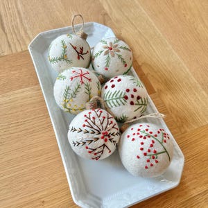Könnte beinhalten: Sechs weiße Filzkugeln mit verschiedenen gestickten Motiven, darunter Schneeflocken, Tannenzweige und Beeren. Die Kugeln sind auf einem rechteckigen weißen Teller angeordnet.