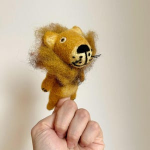 Op de afbeelding: Een vingerpop van vilt van een leeuw met een bruine manen, zwarte ogen en een zwarte neus. De leeuw draagt een geel lichaam en heeft een zwarte mond.