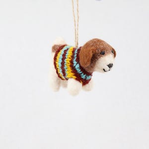 Könnte beinhalten: Ein nadelgefilzter Hundeanhänger mit braunem Kopf und weißem Körper, der einen gestrickten Pullover mit horizontalen Streifen in Gelb, Hellblau und Dunkelrot trägt. Die kleine Figur hängt an einer dünnen, hellbraunen Juteschnur.