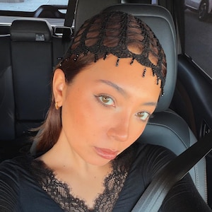 May include: A black long-sleeve top with a lace neckline. The person is wearing a black headpiece with beaded accents. The person is sitting in a car with a seatbelt on.