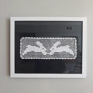 Può includere: Un'opera d'arte in crochet bianco incorniciata con due coniglietti che si fronteggiano. L'opera rettangolare ha un bordo smerlato ed è posta su uno sfondo nero all'interno di una cornice bianca. La cornice è montata su un muro grigio.
