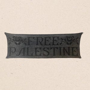 May include: A black rectangular textile with the words "FREE PALESTINE" in capital letters. The text is centered and framed by decorative elements. The background is a light beige color.