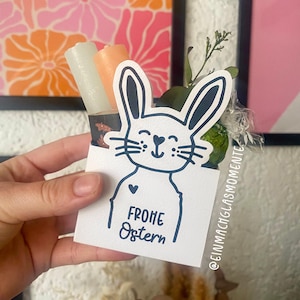 Puede incluir: Una tarjeta blanca con una ilustración de conejo negro y el texto "Frohe Ostern" (Feliz Pascua). La tarjeta se sostiene en una mano y tiene un pequeño corazón dibujado en el pecho del conejo. La tarjeta está decorada con dos velas y una rama de vegetación.