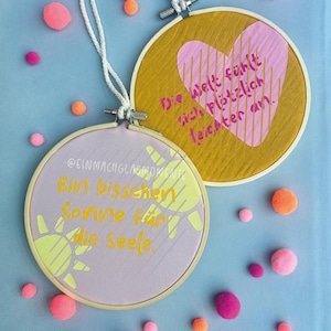 May include: Two round embroidery hoops with text and designs. One has a pink heart on a gold background, the other has a sun design on a lavender background. The hoops are surrounded by pink and orange pom-poms.
