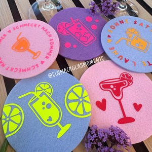 Puede incluir: Un conjunto de posavasos de fieltro de colores, cada uno con un diseño de cóctel diferente en colores neón brillantes. Los diseños incluyen una margarita, un cóctel con hielo y una bebida con pajita. Algunos posavasos tienen texto.