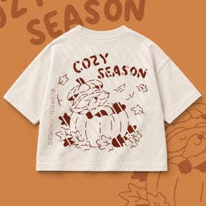 Könnte beinhalten: Ein cremefarbenes T-Shirt mit kurzen Ärmeln und einem dunkelbraunen Aufdruck eines Waschbären, der in einem Kürbis sitzt und von Blättern umgeben ist. Der Text COZY SEASON steht darüber und @EINMACHGLASMOMENTE ist vertikal aufgedruckt.