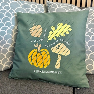 Könnte beinhalten: Ein quadratisches, teal-farbenes Kissen mit einem herbstlichen Motiv. Das Design umfasst einen gelben Kürbis, einen Pilz und ein Blatt mit dem Text "LEAFS ARE FALLING, AUTUMN IS CALLING." Das Kissen trägt auch den Text "@EINMACHGLASMOMENTE."