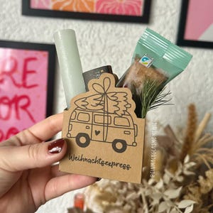 Könnte beinhalten: Eine kleine Geschenkbox aus Kraftpapier mit einer schwarzen Illustration eines Vans, der einen Weihnachtsbaum transportiert, und dem Text "Weihnachtsexpress". Die Box enthält eine hellgrüne Kerze, einen kleinen Schokoriegel und eine verpackte Leckerei.