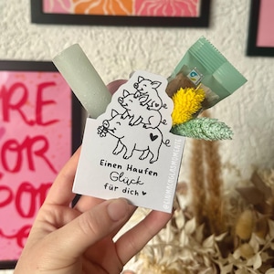 May include: A small gift set held in a hand, featuring a light green cylindrical item, a small packet, and decorative flowers. A white card with a pig illustration and German text is also included.