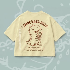 Könnte beinhalten: Ein hellgelbes Kurzarm-T-Shirt mit einer braunen Grafik eines Dinosauriers, der eine Eistüte isst. Der Text lautet SNACKASAURUS und professional ice cream eater, mit dem vertikalen Text @EINMACHGLASMOMENTE auf der linken Seite.