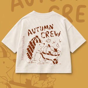 Peut inclure: Un t-shirt à manches courtes blanc cassé avec un motif marron représentant un hérisson portant un bonnet et poussant une brouette remplie d'une citrouille et de feuilles. Le texte "AUTUMN CREW" et "@EINMACHGLASMOMENTE" est imprimé en marron.