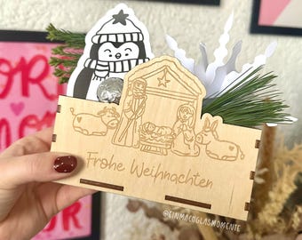 May include: A wooden Christmas box with a laser-etched nativity scene and the words "Frohe Weihnachten." A penguin sticker and greenery are placed inside. The box is held by a person.