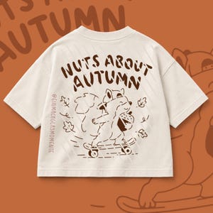 Puede incluir: Una camiseta de manga corta color blanco roto con un gráfico marrón oscuro de una ardilla en un monopatín sosteniendo una bellota. El texto dice NUTS ABOUT AUTUMN y @EINMACHGLASMOMENTE. El diseño incluye hojas y líneas diagonales.