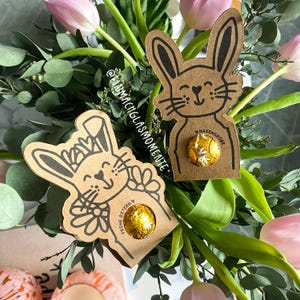 Może przedstawiać: Dwie brązowe, kartonowe etykiety w kształcie królika z czarnymi konturami. Etykiety są ozdobione kwiatami i tekstem "Frohe Ostern" i "Hasengruesse". Każda etykieta ma miejsce na włożenie czekoladowego jajka.