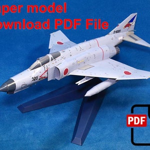 May include: A paper model of a gray and white fighter jet with red markings. The jet is on a blue stand and the text "Paper model Download PDF File" is at the top of the image. A PDF icon with a download arrow is in the lower right corner.