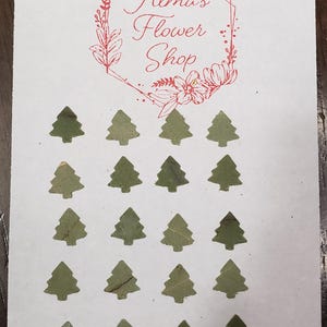 May include: A white card with the text "Alma's Flower Shop" in red lettering inside a wreath design. There are 18 green paper cutouts of Christmas trees on the card.