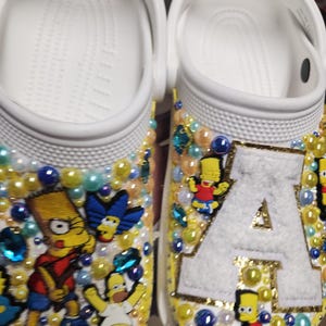 Puede incluir: Zuecos blancos decorados con parches de personajes de dibujos animados, cuentas de colores y pedrería. Los zuecos presentan imágenes de Bart Simpson, Homer Simpson y la letra "A". Los zuecos tienen detalles en amarillo y azul.