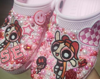 Puede incluir: Zuecos rosas decorados con parches de personajes de dibujos animados, cuentas rosas y blancas y adornos de estrellas y corazones. Los zuecos presentan la palabra "SE" y un botón de carita sonriente. Los zapatos están cubiertos de cuentas rosas, blancas y rojas.