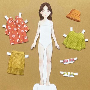 Puede incluir: Mu&ntilde;eca de papel con un cuerpo blanco, cabello casta&ntilde;o y ojos marrones. La mu&ntilde;eca tiene una variedad de opciones de ropa, incluyendo una camisa floral roja y blanca, un su&eacute;ter verde, una falda amarilla, un sombrero marr&oacute;n y zapatos rojos y verdes.