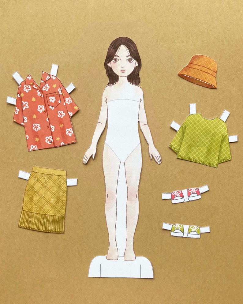 Puede incluir: Mu&ntilde;eca de papel con un cuerpo blanco, cabello casta&ntilde;o y ojos marrones. La mu&ntilde;eca tiene una variedad de opciones de ropa, incluyendo una camisa floral roja y blanca, un su&eacute;ter verde, una falda amarilla, un sombrero marr&oacute;n y zapatos rojos y verdes.