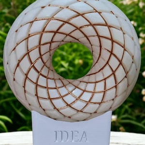 May include: A white circular night light featuring a raised geometric lattice pattern with copper-colored lines. The base is a solid white rectangular block with the word IDEA embossed in capital letters on the front surface of the stand.