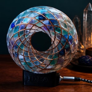 Può includere: Una lampada sferica con un mosaico di tessere di vetro traslucido blu, verdi e viola disposte in cerchio. Un sottile filo di rame è avvolto attorno all'esterno con un motivo a spirale. La lampada poggia su una base nera con la scritta IDEA.