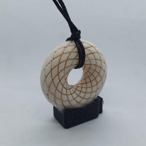 Könnte beinhalten: Eine weiße Halskette mit einem geometrischen Anhänger, bestehend aus einer schwarzen Schnur und einer schwarzen Basis mit der Aufschrift "IDEA".
