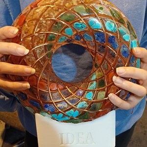 May include: A circular, torus-shaped sculpture featuring a mosaic of multicoloured gemstones embedded in resin, overlaid with a copper wire grid. The sculpture is mounted on a white rectangular base with the word IDEA embossed on the front.