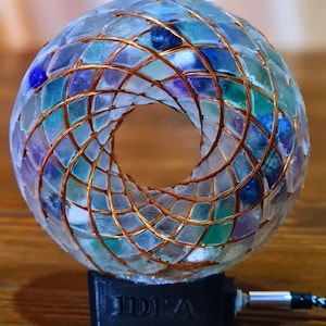 Könnte beinhalten: Eine kreisförmige, durchscheinende Harzskulptur, die mit kleinen, mehrfarbigen Glasfragmenten in Blau-, Grün- und Violetttönen eingebettet ist. Eine Kupferdrahtspule umschließt das Äußere. Die Skulptur steht auf einem schwarzen Sockel mit dem Wort IDEA.