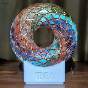 May include: A round, multi-coloured, resin orb with a hole in the centre, decorated with various stones and gold wire. The orb is sitting on a white base with the word "IDEA" embossed on it.
