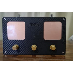 May include: A black electronic device with the word "IDEA" printed on the front. It has two rectangular copper-colored panels and three gold knobs. The device has a carbon fiber texture and two silver connectors.