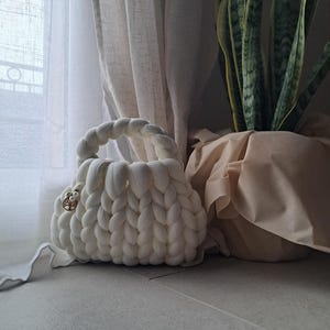 Può includere: Una borsa bianca a maglia grossa con un manico intrecciato e una chiusura decorativa. La borsa è realizzata in filato morbido e spesso e ha una forma arrotondata. Una pianta da interno in un vaso beige è sullo sfondo.