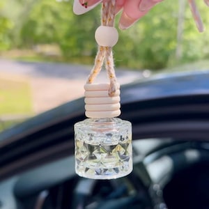 May include: A clear, faceted glass car air freshener with a wooden top and a beige rope for hanging. The air freshener is filled with a clear liquid and has a decorative wooden bead. The background is blurred.