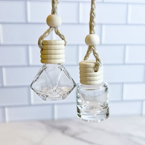 May include: Two glass car air freshener bottles with wooden caps and string hangers. The bottle on the left is a diamond shape and the bottle on the right is round.