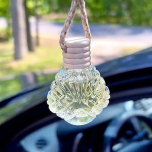 May include: A clear, decorative car air freshener with a floral-patterned glass bottle filled with yellow liquid. It hangs from a braided rope with a wooden bead. The car's interior and a blurred green background are visible.