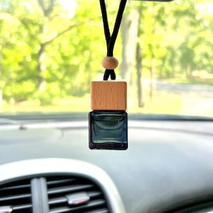 May include: A car air freshener with a wooden top and a square glass bottle filled with blue liquid. The freshener hangs from a black cord with a wooden bead, suspended in front of a car's dashboard and windshield.
