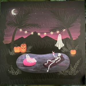 May include: A skeleton wearing a swimsuit relaxes in a pool with a pink cocktail. Two ghosts float in the background, one on a swing and one in the pool. Palm trees and jack-o-lanterns decorate the scene.