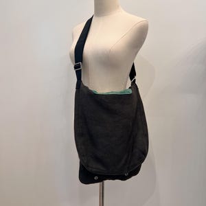 Puede incluir: Un bolso mensajero gris oscuro con una correa negra se exhibe en un maniqu&iacute;. El bolso tiene un cierre de solapa con dos cierres a presi&oacute;n. El forro interior es de color verde claro. El bolso est&aacute; hecho de una tela texturizada.
