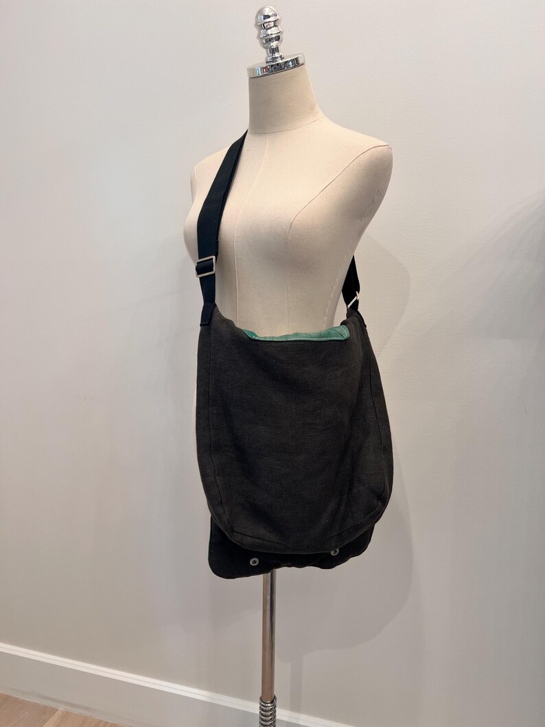 Puede incluir: Un bolso mensajero gris oscuro con una correa negra se exhibe en un maniqu&iacute;. El bolso tiene un cierre de solapa con dos cierres a presi&oacute;n. El forro interior es de color verde claro. El bolso est&aacute; hecho de una tela texturizada.
