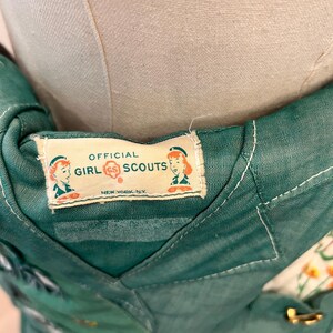 Puede incluir: Primer plano de un uniforme vintage de Girl Scouts. La tela verde tiene una etiqueta que dice "Official Girl Scouts New York, NY". El uniforme tiene varios parches y un cierre dorado.