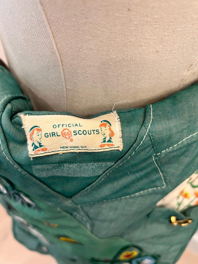 Puede incluir: Primer plano de un uniforme vintage de Girl Scouts. La tela verde tiene una etiqueta que dice "Official Girl Scouts New York, NY". El uniforme tiene varios parches y un cierre dorado.
