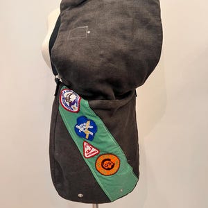 Puede incluir: Un bolso mensajero de lona gris oscuro con una banda verde con varios parches. El bolso tiene una parte superior redondeada y correas ajustables. Los parches incluyen texto y gr&aacute;ficos. El bolso se exhibe en un maniqu&iacute;.