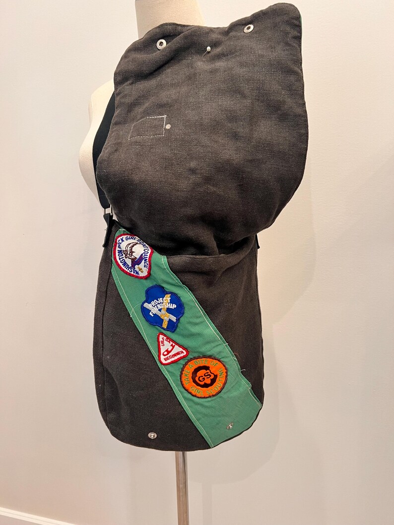 Puede incluir: Un bolso mensajero de lona gris oscuro con una banda verde con varios parches. El bolso tiene una parte superior redondeada y correas ajustables. Los parches incluyen texto y gr&aacute;ficos. El bolso se exhibe en un maniqu&iacute;.