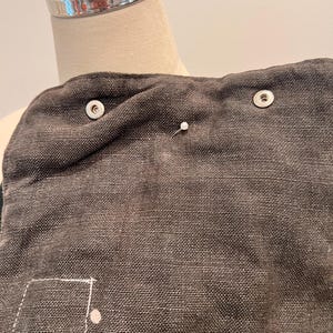 Puede incluir: Primer plano de una tela gris oscuro con un tejido texturizado. Se ven dos broches plateados, junto con un alfiler blanco y un cuadrado cosido con un punto. La tela parece ser parte de una prenda o accesorio.
