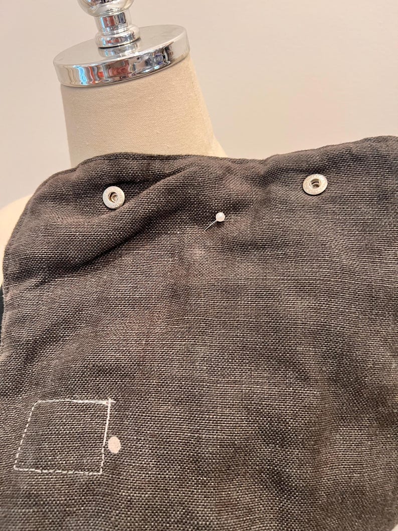 Puede incluir: Primer plano de una tela gris oscuro con un tejido texturizado. Se ven dos broches plateados, junto con un alfiler blanco y un cuadrado cosido con un punto. La tela parece ser parte de una prenda o accesorio.