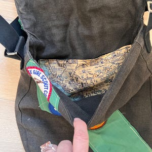 Puede incluir: Bolso mensajero de lona gris oscuro con correa negra. El forro interior presenta un dise&ntilde;o de mapa. Los parches incluyen un logotipo de Girl Scout y una solapa verde con cierre de bot&oacute;n a presi&oacute;n. El bolso est&aacute; abierto.