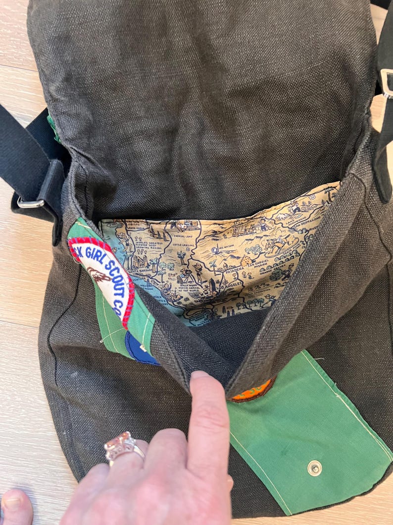 Puede incluir: Bolso mensajero de lona gris oscuro con correa negra. El forro interior presenta un dise&ntilde;o de mapa. Los parches incluyen un logotipo de Girl Scout y una solapa verde con cierre de bot&oacute;n a presi&oacute;n. El bolso est&aacute; abierto.