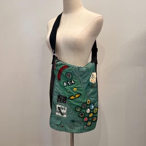 Puede incluir: Un bolso de hombro verde hecho de una banda de Girl Scout. El bolso presenta varios parches, incluyendo el n&uacute;mero "814" y las palabras "Girl Scout". El bolso tiene una correa negra y se muestra en un maniqu&iacute;.