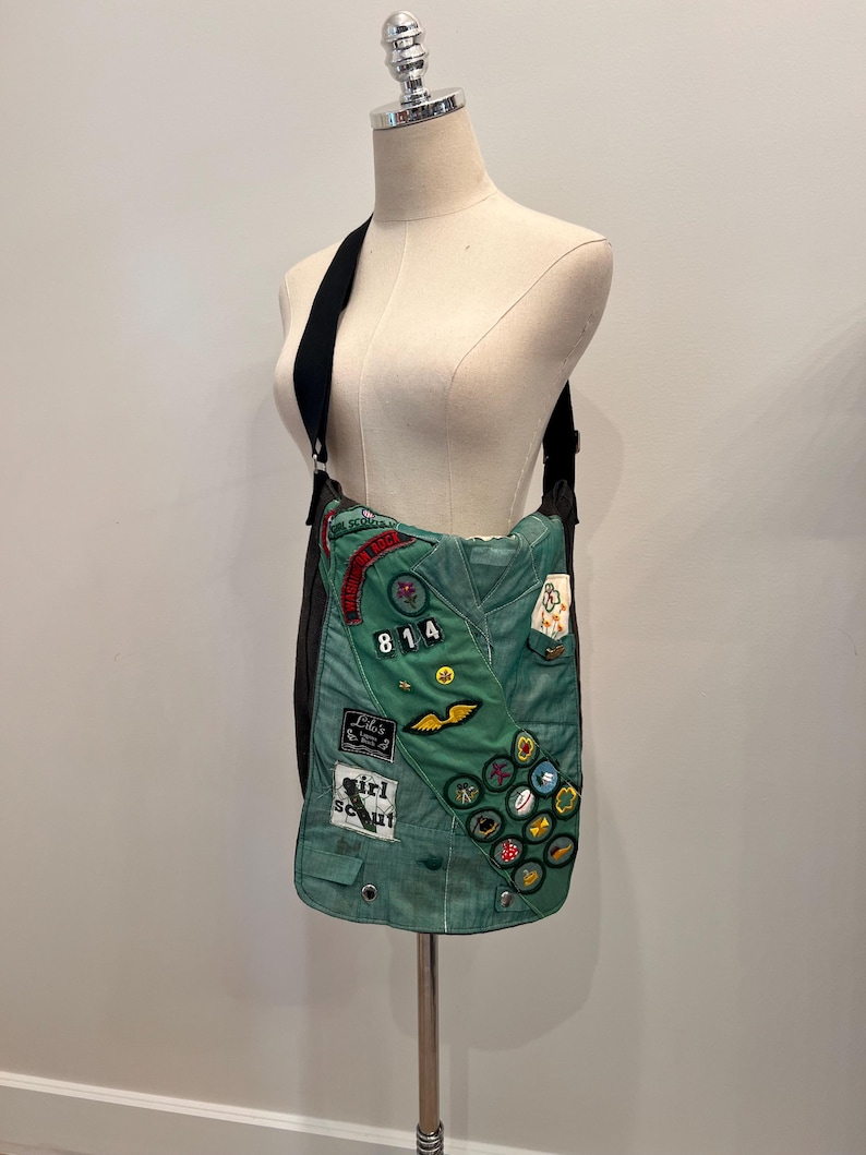 Puede incluir: Un bolso de hombro verde hecho de una banda de Girl Scout. El bolso presenta varios parches, incluyendo el n&uacute;mero "814" y las palabras "Girl Scout". El bolso tiene una correa negra y se muestra en un maniqu&iacute;.