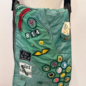 Puede incluir: Un bolso de hombro verde hecho de un uniforme de Girl Scout. El bolso presenta varios parches bordados, incluyendo insignias, un n&uacute;mero de unidad y una etiqueta con el nombre. Tiene una correa negra y un respaldo gris.
